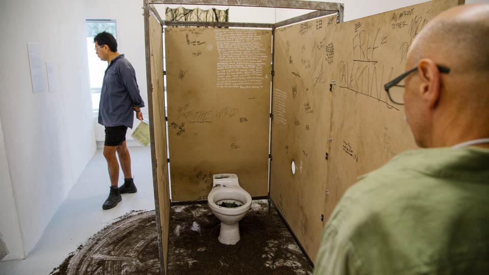 Graffiti covered cubicle set on earth-scattered space in gallery. Two males viewing the piece. 