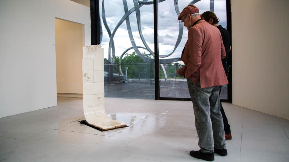 Two males view a urinal line structural sculpture, with taps set atop it, running water streaming down sculpture. 