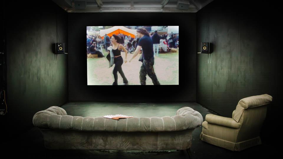 Video projection showing a couple, one male,one female. In front of the wall on which the video is projected is a cream-coloured settee and armchair. 
