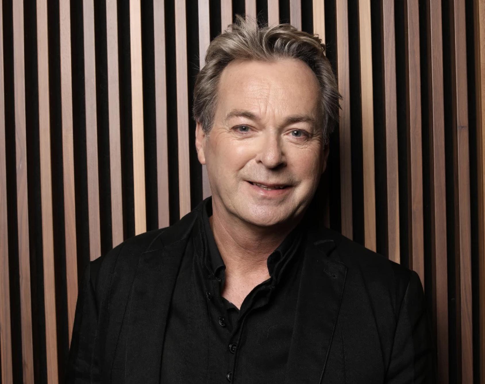 A portrait photograph of Julian Clary, smiling as he wears a black shirt and jacket