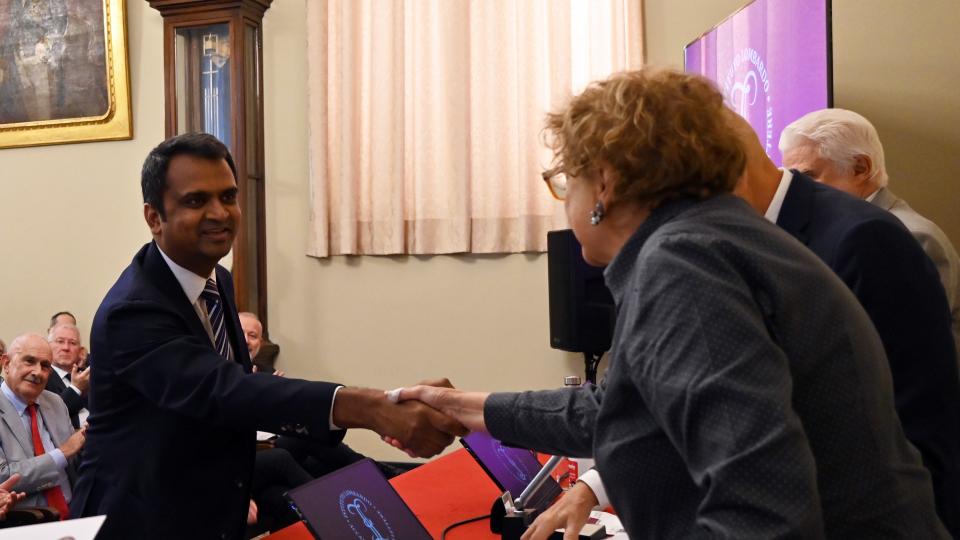 Professor Ragupathy Venkatachalam, wearing a suit and tie, shakes hands with a woman. 